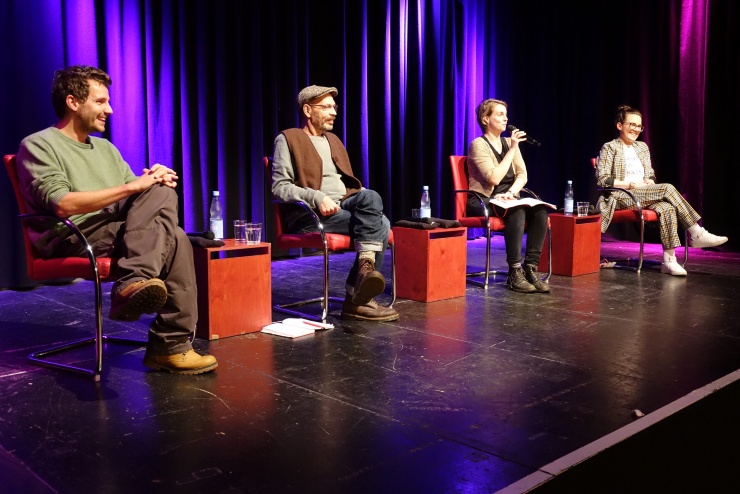 Vier Menschen sitzen auf Stühlen auf einer bunt ausgeleuchteten Bühne