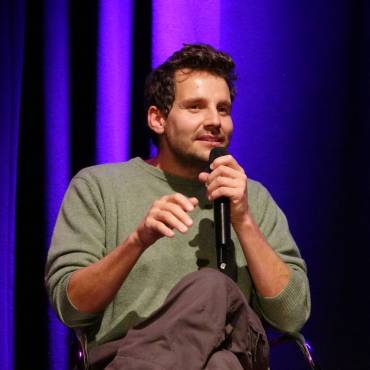 Der Autor Tobi Rosswog spricht in ein Mikrofon, im Hintergrund ein Bühnenvorhang