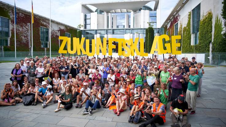 Eine große Gruppe Menschen sitzt und steht vor dem Kanzleramt, sie halten Buchstaben an Stäben hoch, die das Wort ZUKUNFTSKLAGE ergeben