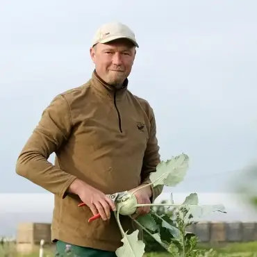 Vor einem Hof ein Landwirt in brauner Jacke, er hält einen Kohlrabi in der Hand.