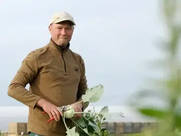 Vor einem Hof ein Landwirt in brauner Jacke, er hält einen Kohlrabi in der Hand.