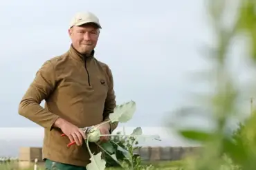 Vor einem Hof ein Landwirt in brauner Jacke, er hält einen Kohlrabi in der Hand.