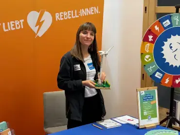 Eine junge Frau steht lächelnd hinter einem Messetisch der EWS, sie hat ein kleines Modell eines Windkraftwerks in der Hand.
