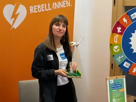 Eine junge Frau steht lächelnd hinter einem Messetisch der EWS, sie hat ein kleines Modell eines Windkraftwerks in der Hand.