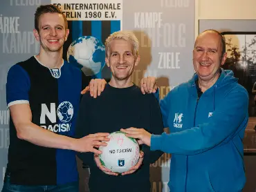 Drei junge Männer legen vor einer Wand mit Sporvereinslogo ihre Hände auf einen Fußball, auf dem «No Racism» steht.