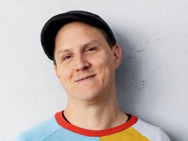Portrait des Kabarettisten Sebastian 23 in buntgemustertem Pullover vor einer grauen Wand