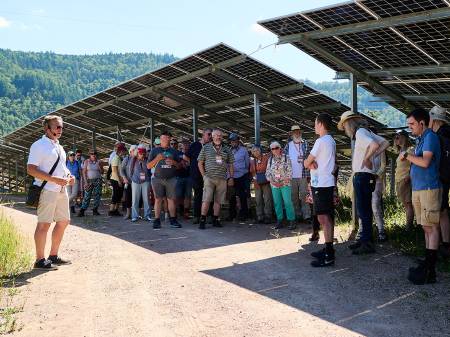 Eine große Gruppe von Menschen hat sich an einem Sommertag in den Schatten der PV-Module eines Solarparks geflüchtet, nur ein Guide steht davor auf dem Weg
