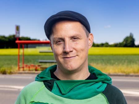 Portrait des Kabarettisten Sebastian 23 in buntgemustertem Pullover vor einer grauen Wand