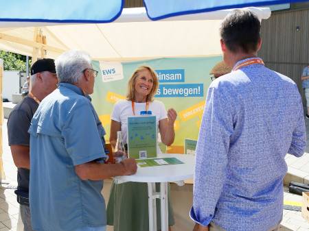 An einem Marktstand steht eine lächelnde Frau, im Hintergrund ist ein Banner der EWS angebracht. Davor sind mehrere Männer von hinten zu sehen, die mit der Frau diskutieren.