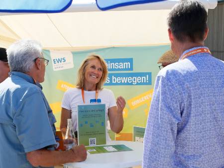 An einem Marktstand steht eine lächelnde Frau, im Hintergrund ist ein Banner der EWS angebracht. Davor sind mehrere Männer von hinten zu sehen, die mit der Frau diskutieren.