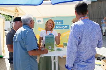 An einem Marktstand steht eine lächelnde Frau, im Hintergrund ist ein Banner der EWS angebracht. Davor sind mehrere Männer von hinten zu sehen, die mit der Frau diskutieren.