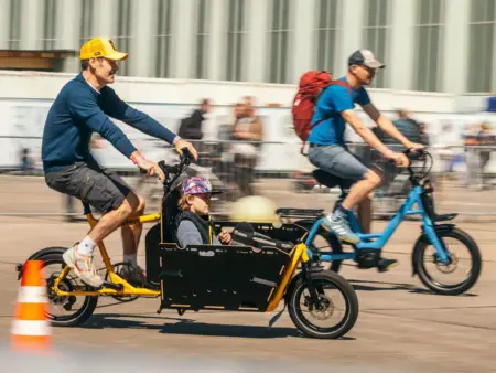 Auf einer Rennstrecke testen Menschen Fahrräder, zentral im Bild ist ein Vater mit einem Lastenrad, in dessen Ladefläche ein Kind sitzt
