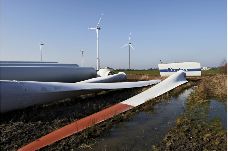 Unmotiert liegen Teile einer Windkraftlage auf einer Wiese, im Hintergrund ragen mehrere Windkraftanlagen empor.