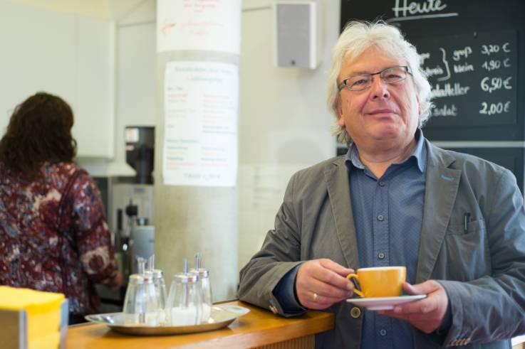 Ein lächelnder älterer Herr steht mit einer Tasse in einer Cafeteria.