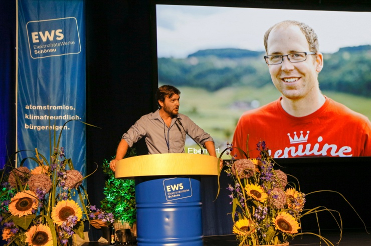 Sebastian Sladek auf der Bühne und hält die Laudatio auf den Stromrebellen Syril Eberhardt, dessen Foto hinter ihm auf der Leinwand erscheint.