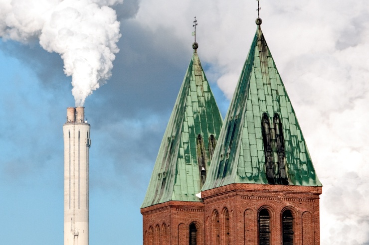 Zwei Kirchturmspitzen davor ein hoher qualmender Schornstein.