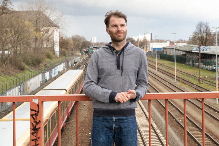 Ein junger Mann steht mit locker verschränkten Armen auf einer Brücke, die über Gleise führt.