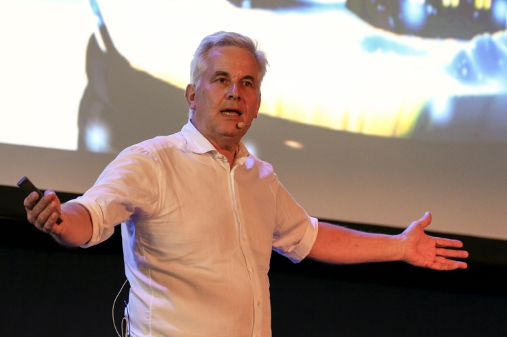 Ein älterer Mann mit grauen Haaren steht vor einer Videoleinwand und breitet seine Arme aus.