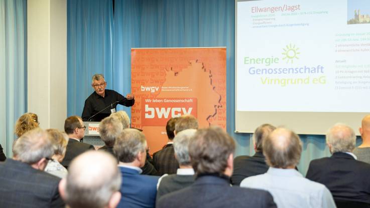 Blick in den gefüllten Veranstaltungsaal Richtung Bühne. Ein Mann steht am Podium und präsentiert ein Projekt.