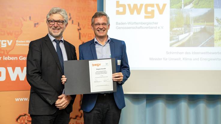 Zwei Männer stehen nebeneinander auf der Bühne. Einer präsentiert eine Urkunde.
