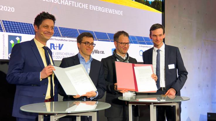 Vier Herren auf einer Bühne präsentieren unterzeichnete Dokumente dem Publikum.