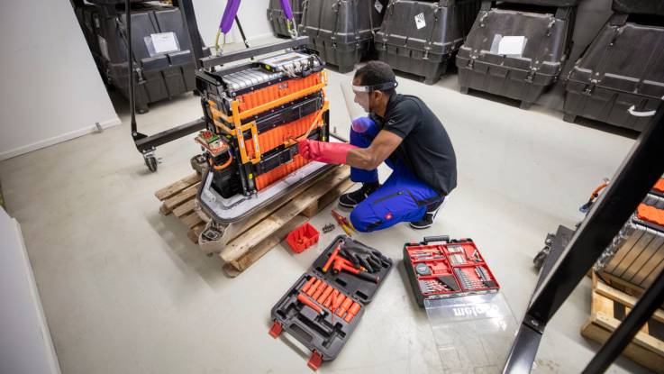Eine etwa ein Meter hohes Batterie-Modul steht auf einer Palette in einer sehr sauberen Werkhalle. Ein Mechaniker arbeitet daran mit Schutzhandschuhen.