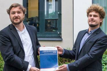 Zwei Männer in Anzug und strubbeliger Frisur halten ein Papierdokument freudig und stolz hoch.