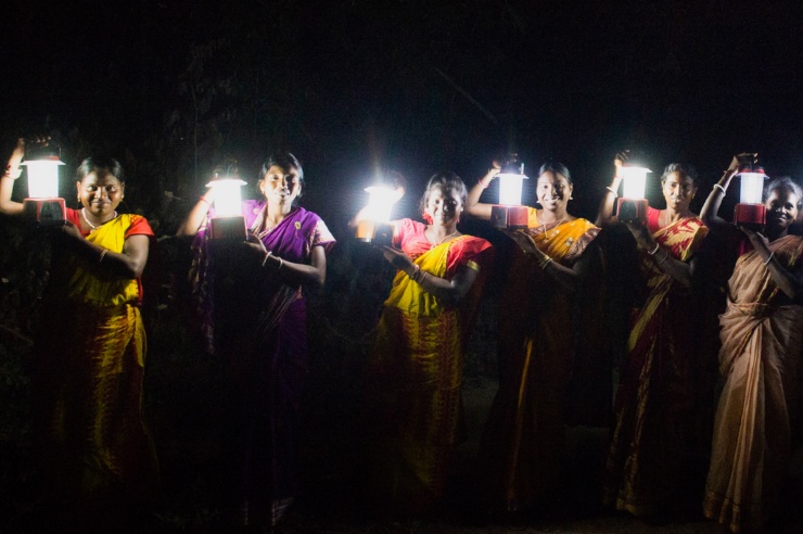 In der Dunkelheit steht eine Reihe indischer Frauen und Männer. Jede hält eine strahlende Lampe in die Höhe. 