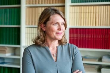 Eine Frau steht vor einem Bücherregal mit zahlreichen gleich aussehenden Büchern und schaut Richtung Fenster