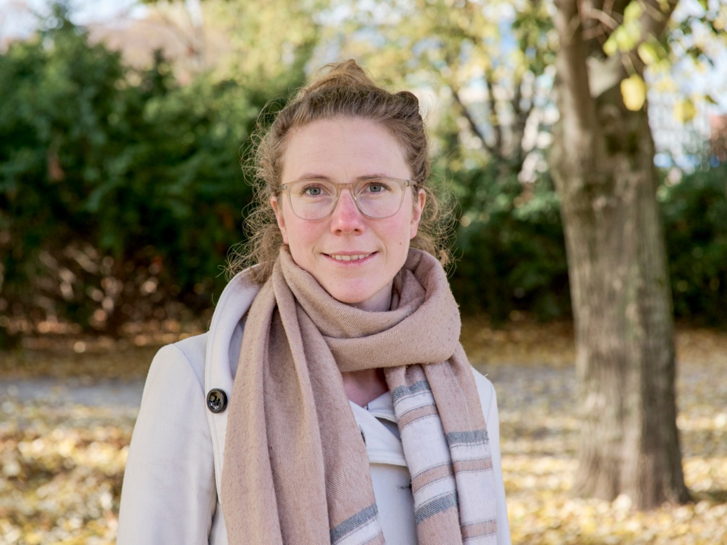 A friendly-looking young woman stands in front of autumnal trees and smiles into the camera.
