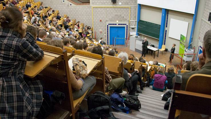 View down the steps of a full auditorium. An older man stands in the stage area, addressing the audience.