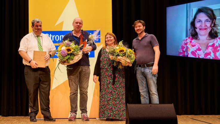 Vier Personen, die zwei in der Mitte mit Trophäe und Blumen im Arm, stehen auf der Bühne, im Hintergrund rechts ein Monitor, auf dem eine lächelnde Frau zu sehen ist.auf dem Monitor ist 