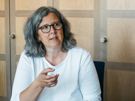 Eine Frau mit schulterlangem, grau-meliertem Haar und Brille schaut interessiert, während sie eine Hand hebt.