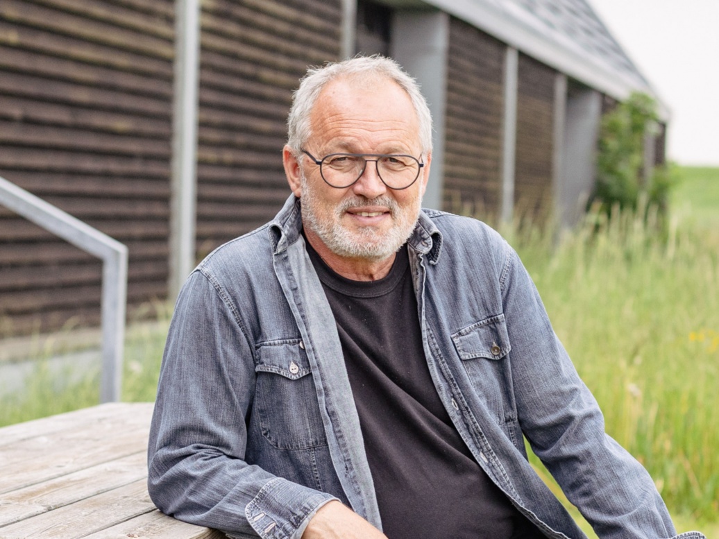 Ein Mann mit kurzem grauem Haar, Brille und ausgewaschenem Jeanshemd sitzt lächelnd im Freien.