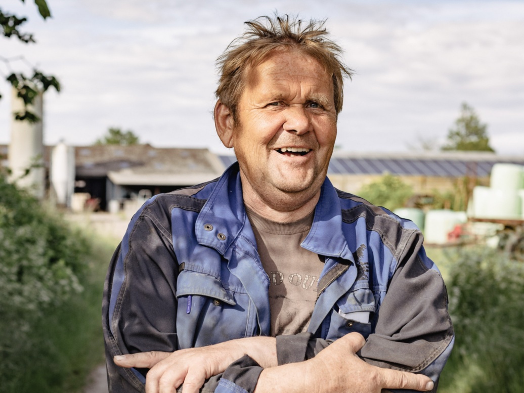 Ein Mann mit rundem Gesicht, blondem Haar und kräftigen Händen steht mit verschränkten Armen im Freien und lacht.
