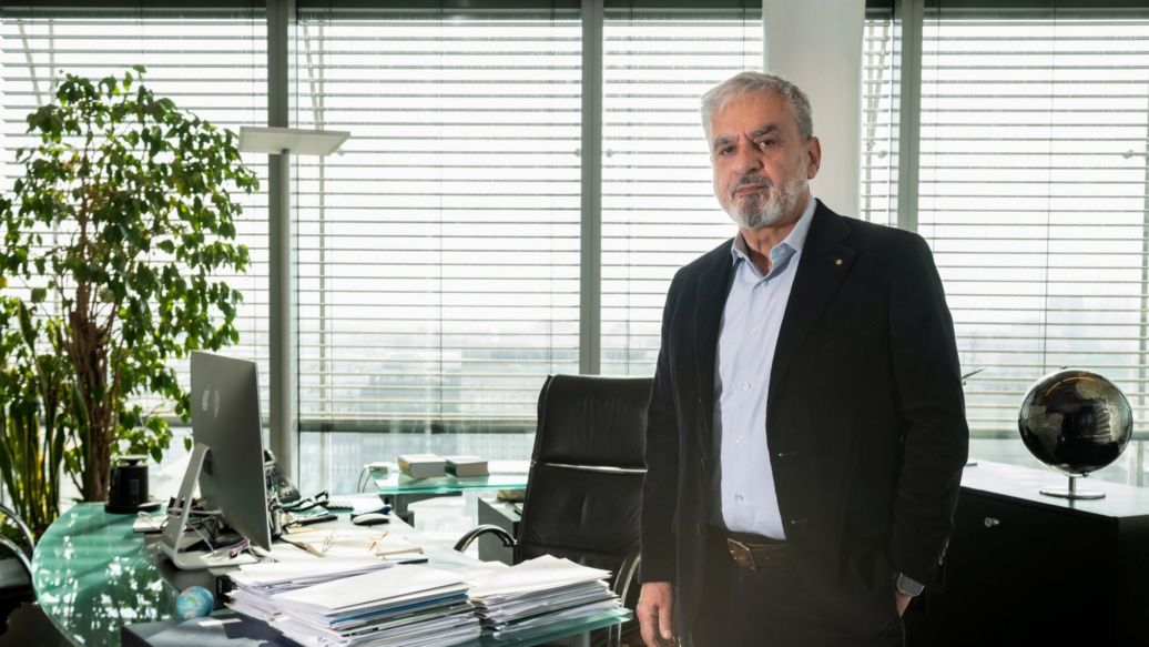 Ein Mann im Anzug mit grauem Haar und Bart steht im Büro – auf dem Schreibtisch neben ihm stapeln sich Unterlagen. 