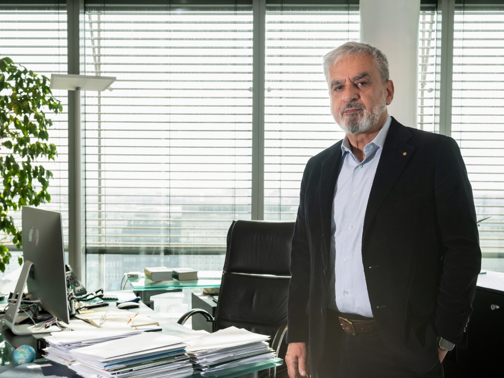 A man in a suit with grey hair and a beard is standing in the office - documents are piled up on the desk next to him. 