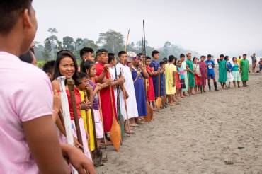 Zahlreiche Menschen stehen bunt gekleidet im Halbkreis am Ufer eines Flusses und halten Ruder in den Händen.