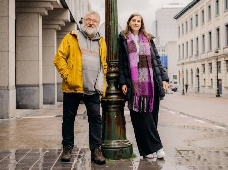  Ein Mann mit grauem Haar und eine jüngere Frau mit hellbraunem Haar lehnen sich an einer grünen Laterne an. 