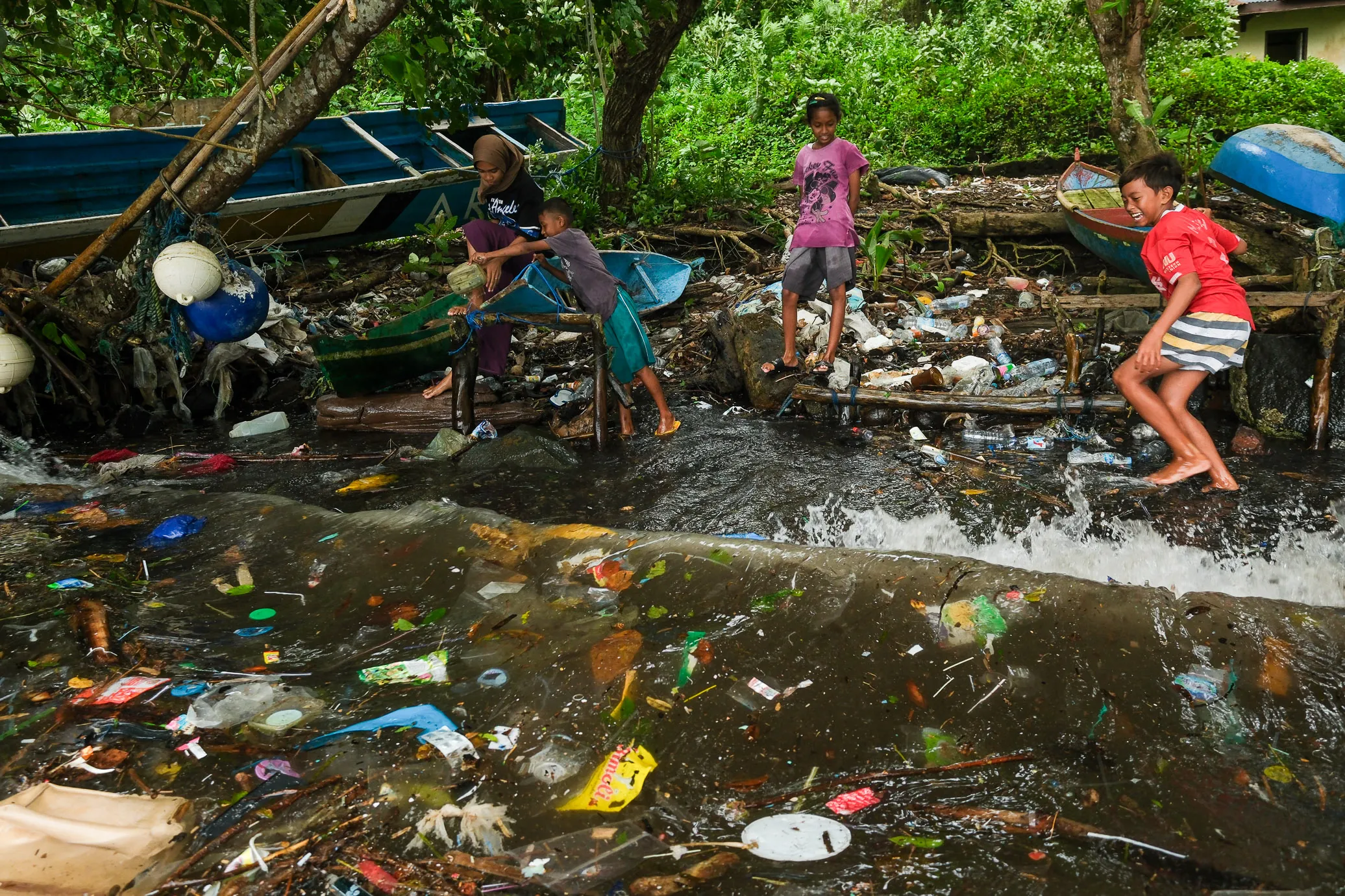 Ein Gruppe von Kindern spielt im Wasser einer steinigen Brandung – überall um sie herum befindet sich Plastikmüll. 