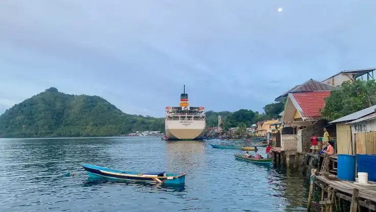 In einer Bucht reihen sich zahlreiche Hütten aneinander, im Wasser liegt ein Fährschiff vor Anker.