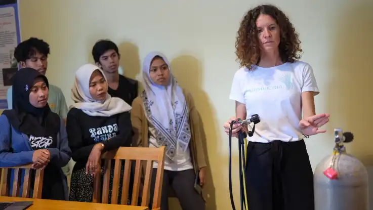 Student:innen hören einer Frau mit braunen Locken zu. Diese deutet auf eine Sauerstoffflasche, während sie in der anderen Hand einen Atemregler hält.