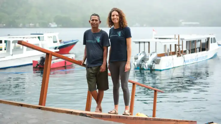 Ein Mann und eine Frau stehen lächeln auf einem Landungssteg, hinter ihnen treiben einige Boote im ruhigen Wasser der Bucht. 