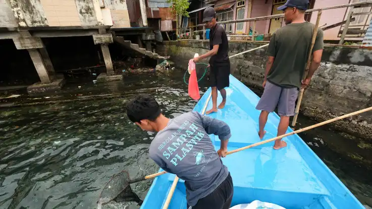 Drei junge Männer fischen mit Köchern von einem blauen Boot aus Plastikmüll aus dem Wasser. 