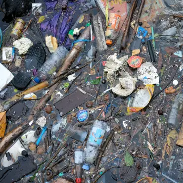 Auf der Wasseroberfläche eines trüben Gewässers schwimmen zahlreiche Alltagsgegenstände und Verpackungen aus Plastik. 