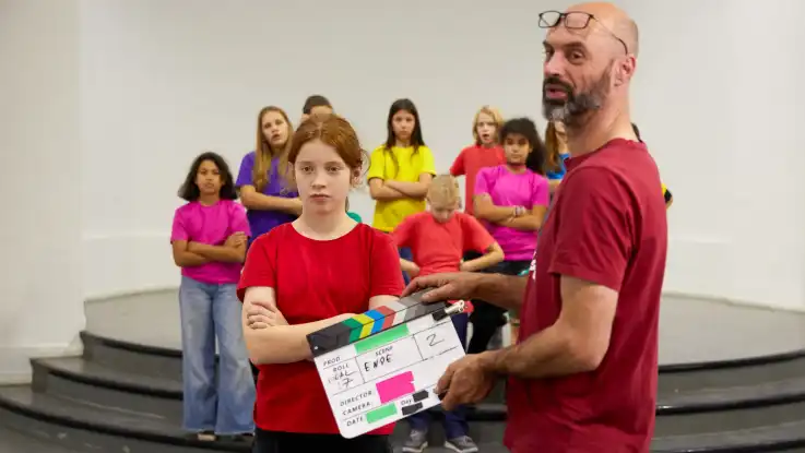 Ein Mann mit Glatze hält eine Filmklappe in den Händen – dahinter posieren die zwölf Kinder mit verschränkten Armen. 