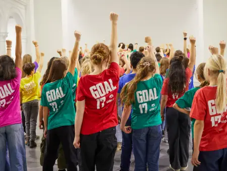 Dutzende Kinder mit bunten T-Shirts stehen in einem weißen Saal und recken die rechte Faust solidarisch nach oben. 