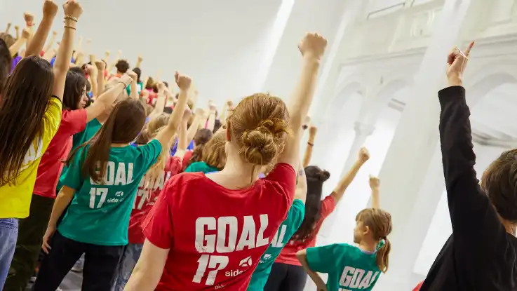 Dutzende Kinder mit bunten T-Shirts stehen in einem weißen Saal und recken die rechte Faust solidarisch nach oben. 