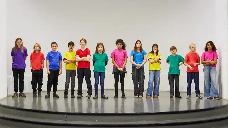 Zwölf Kinder stehen in bunten T-Shirts auf einem Podest, die Blicke in Richtung der Kamera gerichtet.