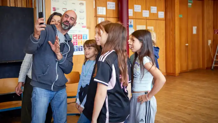 Der Mann mit der Glatze zeigt den Kindern etwas auf seinem Smartphone, eine Frau schaut ihm dabei über die Schulter. 